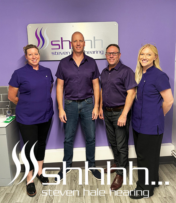 Steven Hale Hearing team standing together in the Sutton Coldfield clinic, wearing purple uniforms in front of the clinic sign.